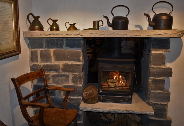 A stone fireplace with a wood-burning stove, a mantelpiece with various metal pots and a kettle, and a wooden chair.
