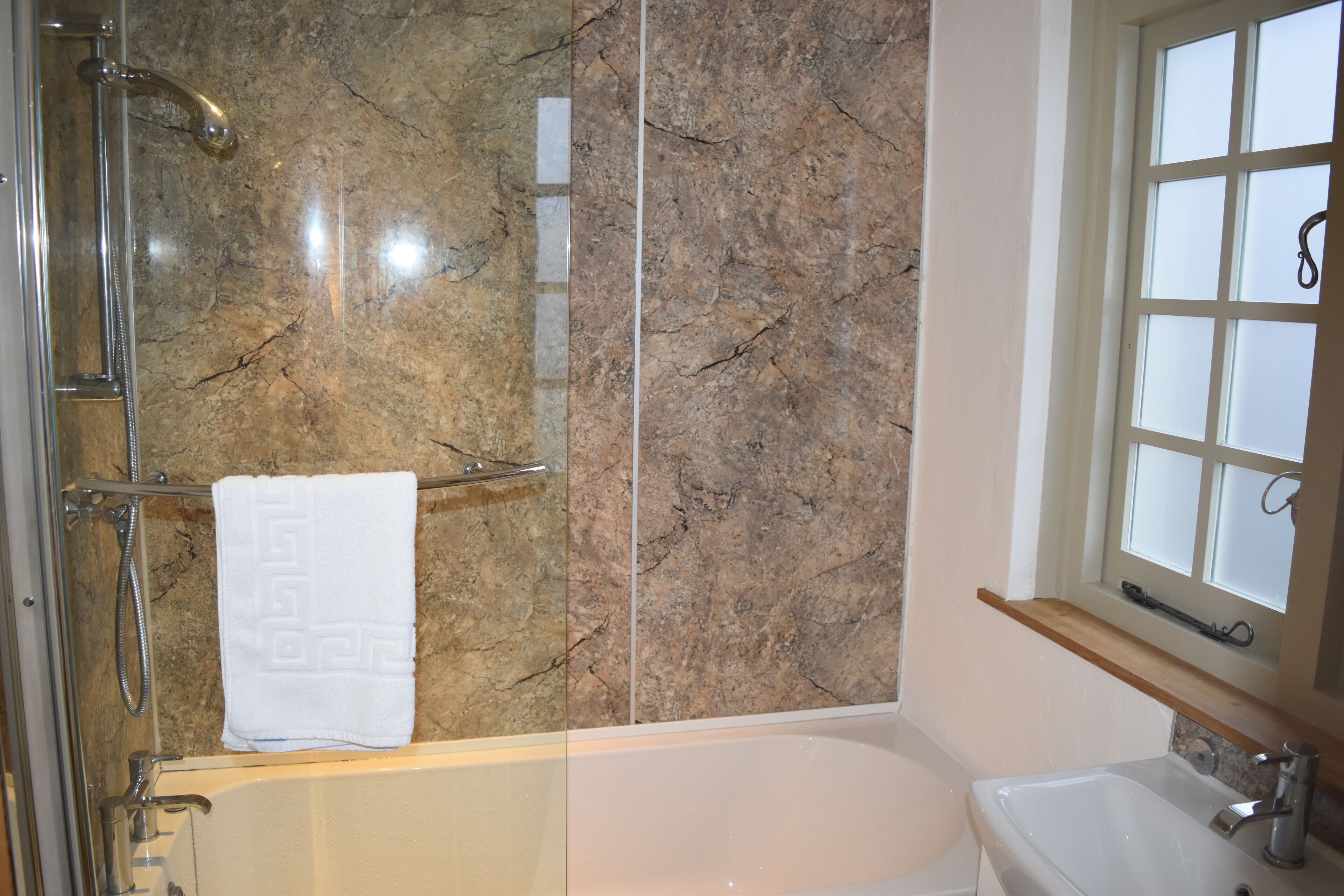 A bathroom with a marble wall, a bathtub, a sink, and a window with frosted glass.