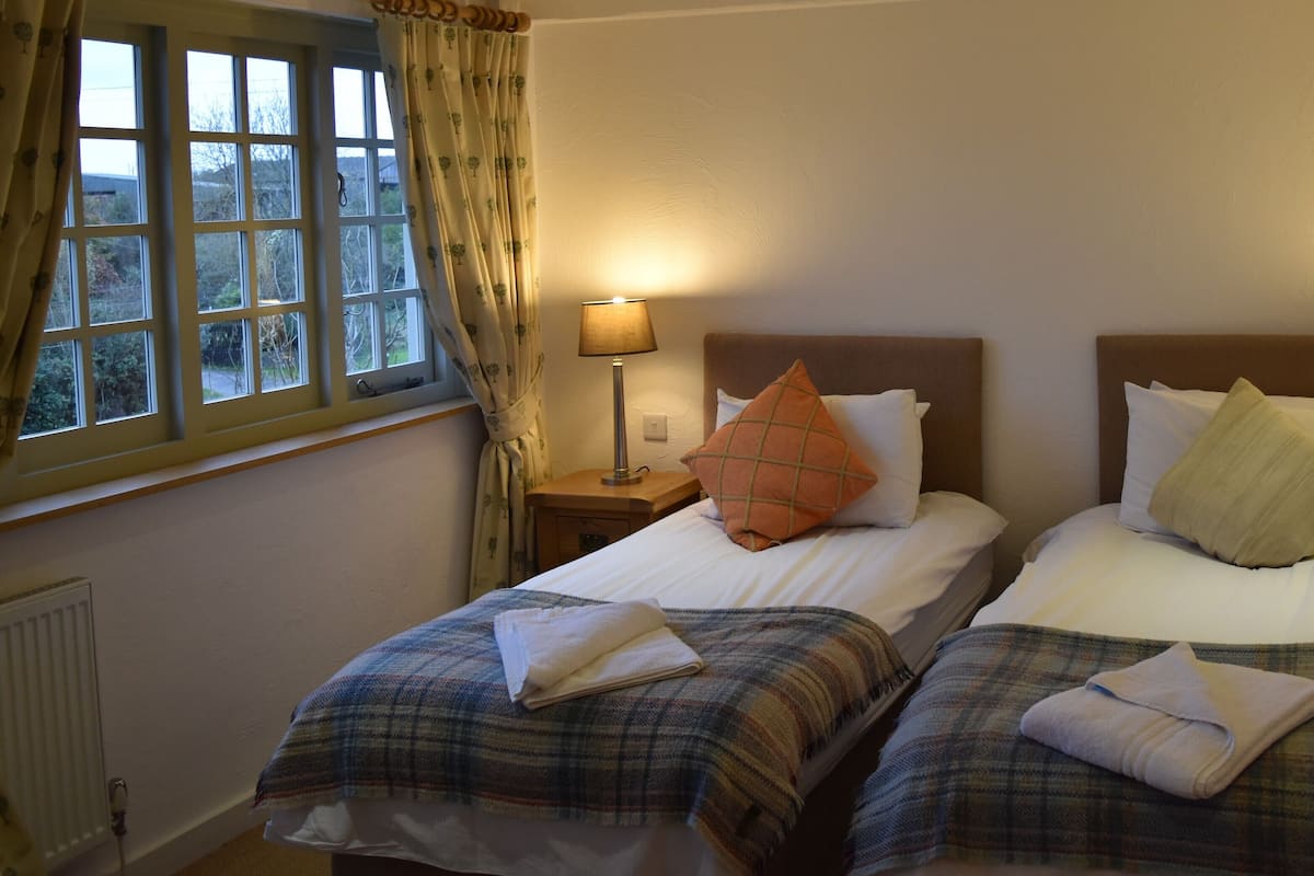 Quarto de hotel com duas camas, cada uma com cabeceira, mesas de cabeceira e luminárias. Há uma janela com cortinas no lado esquerdo.