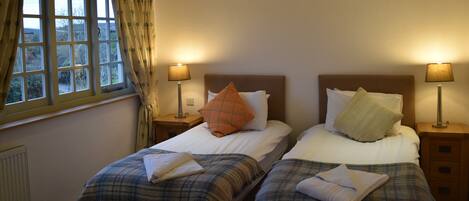 A hotel room with two beds, each with a headboard, bedside tables, and lamps. There is a window with curtains on the left side.