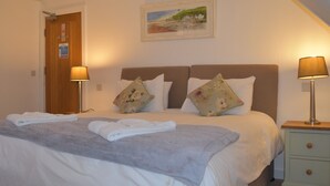 A neatly made bed with a grey blanket and two pillows, a wooden door with a sign, a framed picture on the wall, and two bedside lamps. - Three Horseshoes (Barry)