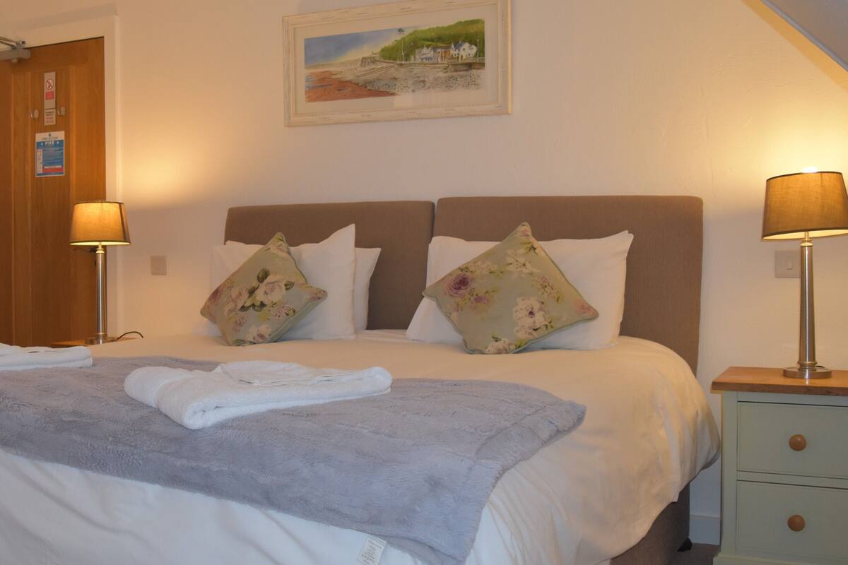 Uma cama bem arrumada com um cobertor cinza e duas almofadas, uma porta de madeira com uma placa, um quadro na parede e duas luminárias de mesa.