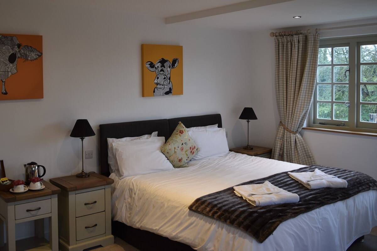 Cama bem arrumada com lençóis brancos, duas luminárias de cabeceira, mesa de cabeceira com abajur, pequena mesa com chaleira e xícaras, e obras de arte nas paredes.