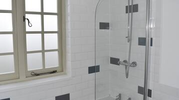 Ein Badezimmer mit einer gläsernen Duschkabine, einem Fenster mit gebürstetem Glas und weißen Fliesenwänden mit schwarzen Akzenten.