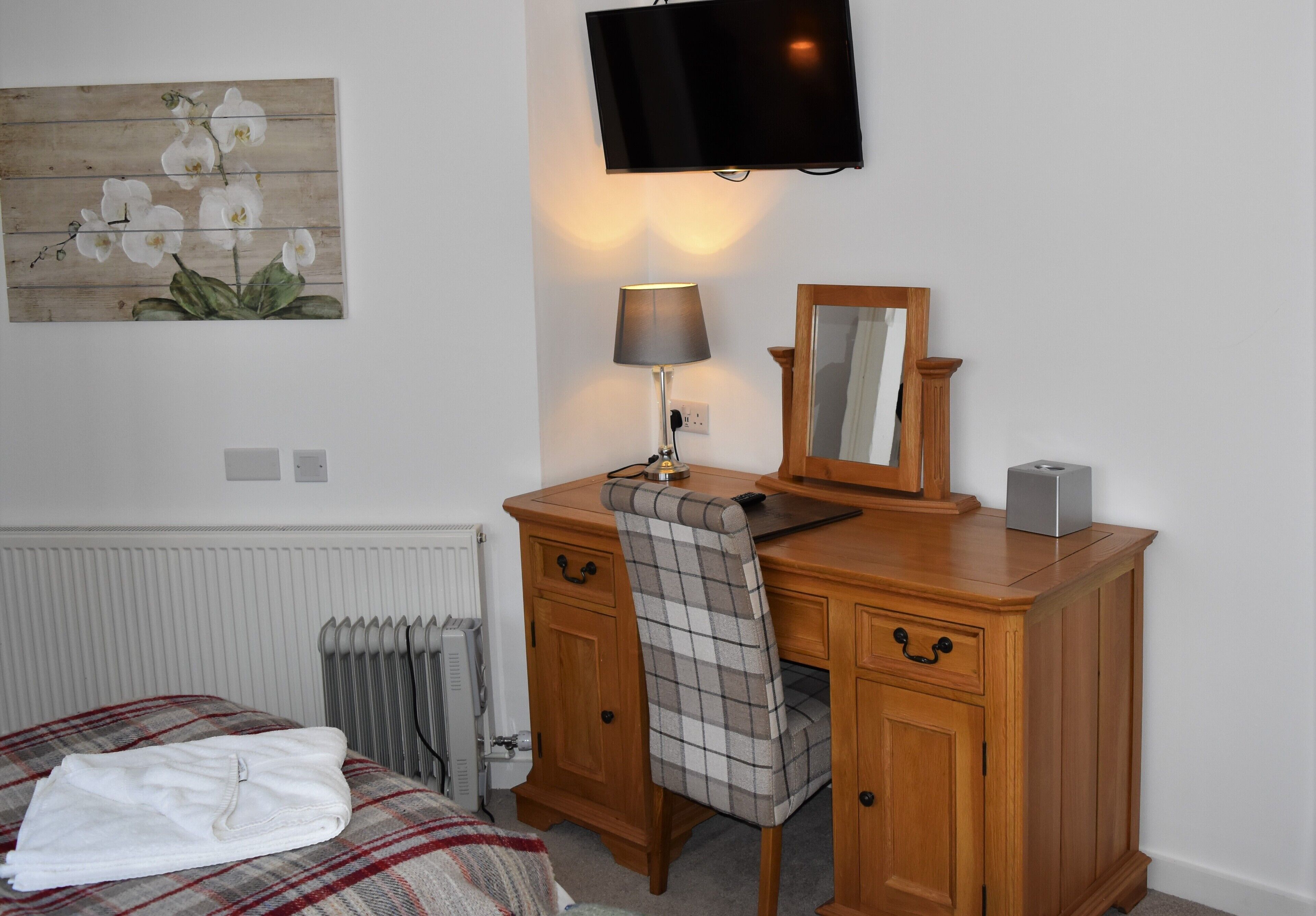A hotel room with a wooden desk, a chair, a television, a lamp, a mirror, and a bed with a plaid blanket.