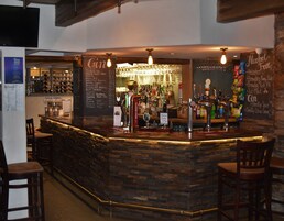 Uma área de bar com bancada de pedra, diversas torneiras e variedade de garrafas e copos.