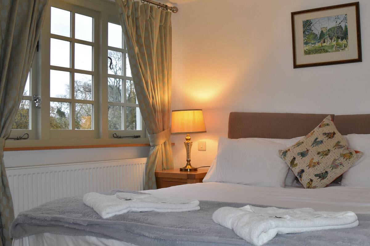 Quarto de hotel com cama, mesa de cabeceira, abajur, janela com cortinas e um quadro na parede.