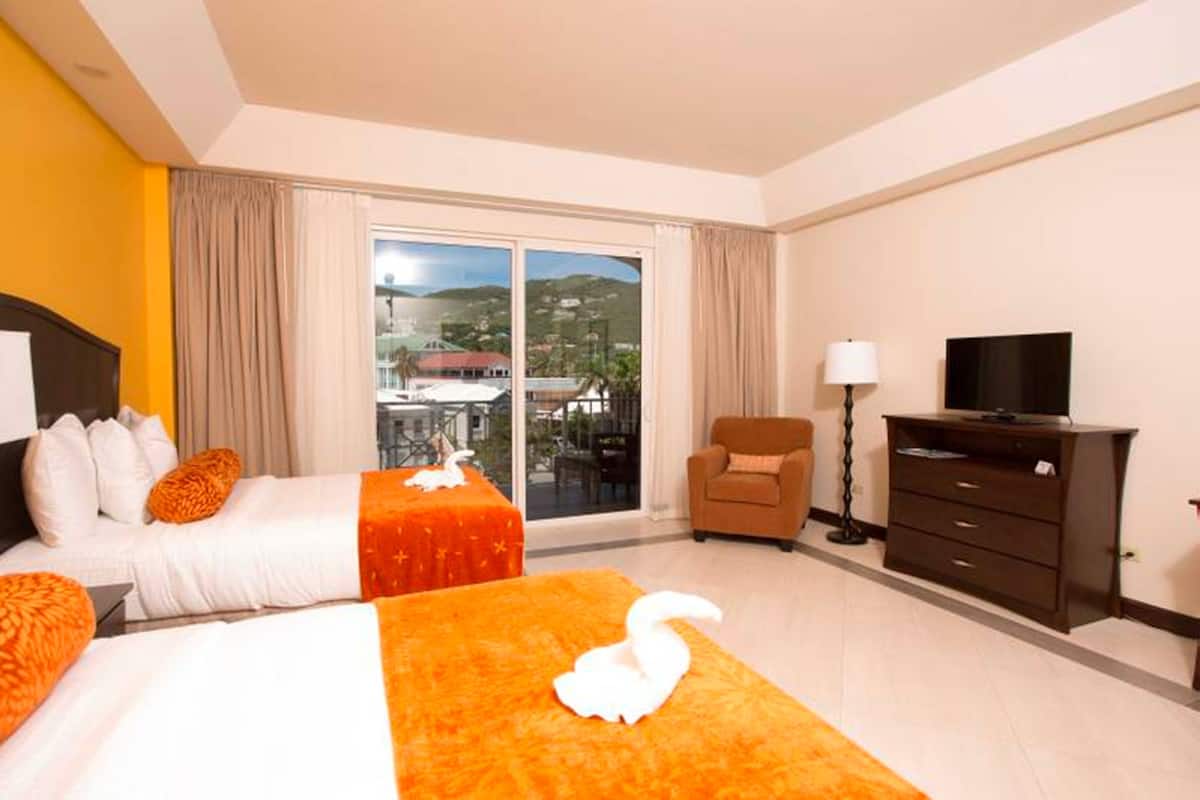 Une chambre d’hôtel avec deux lits, une télévision, une chaise et une grande fenêtre donnant sur une ville et des collines.