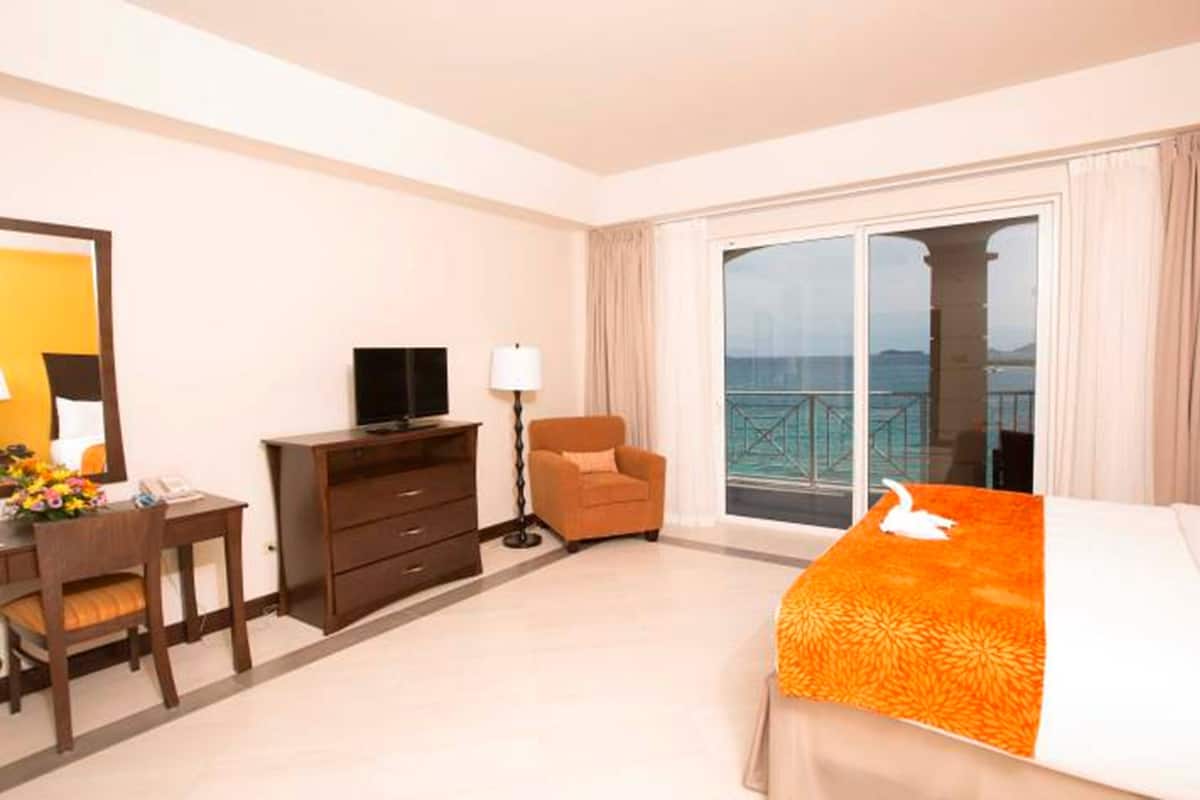 Une chambre d’hôtel comprenant un lit, un bureau avec une chaise, une télévision, une lampe et une vue sur la mer grâce à de grandes fenêtres.