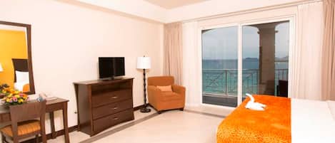 A hotel room with a bed, a desk with a chair, a television, a lamp, and a view of the sea through large windows.