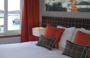 A hotel room with a bed, pillows, a nightstand, a lamp, a window with a view of boats, and a painting on the wall. - Anchor Hotel (Tarbert)