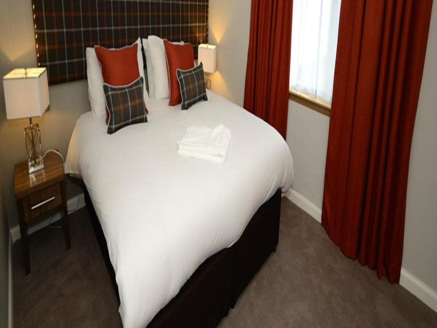 A neatly made bed with white linens and plaid pillows, a bedside table with a lamp, and red curtains.