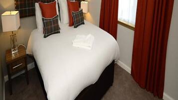 A neatly made bed with white linens and plaid pillows, a bedside table with a lamp, and red curtains.