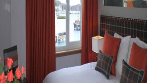 A hotel room with a bed, red curtains, a window with a view of a body of water and boats, and a painting on the wall.