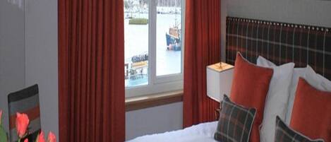 A hotel room with a bed, red curtains, a window with a view of a body of water and boats, and a painting on the wall.