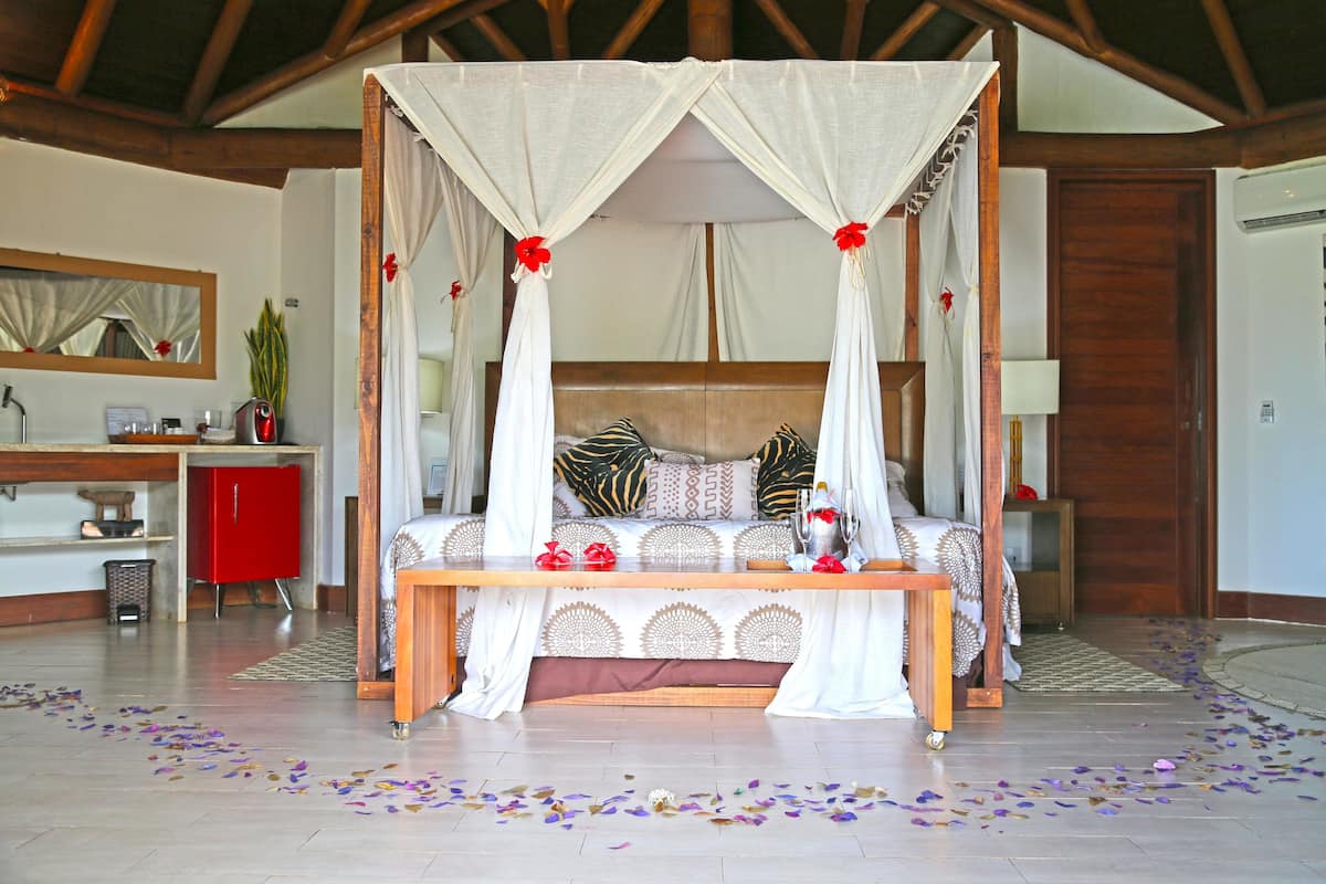 Un lit à baldaquin à quatre poteaux avec une tête de lit en bois, une table de chevet ornée d’un vase et d’une fleur rouge, et une armoire rouge dans une pièce dont le plafond est décoré de poutres en bois.