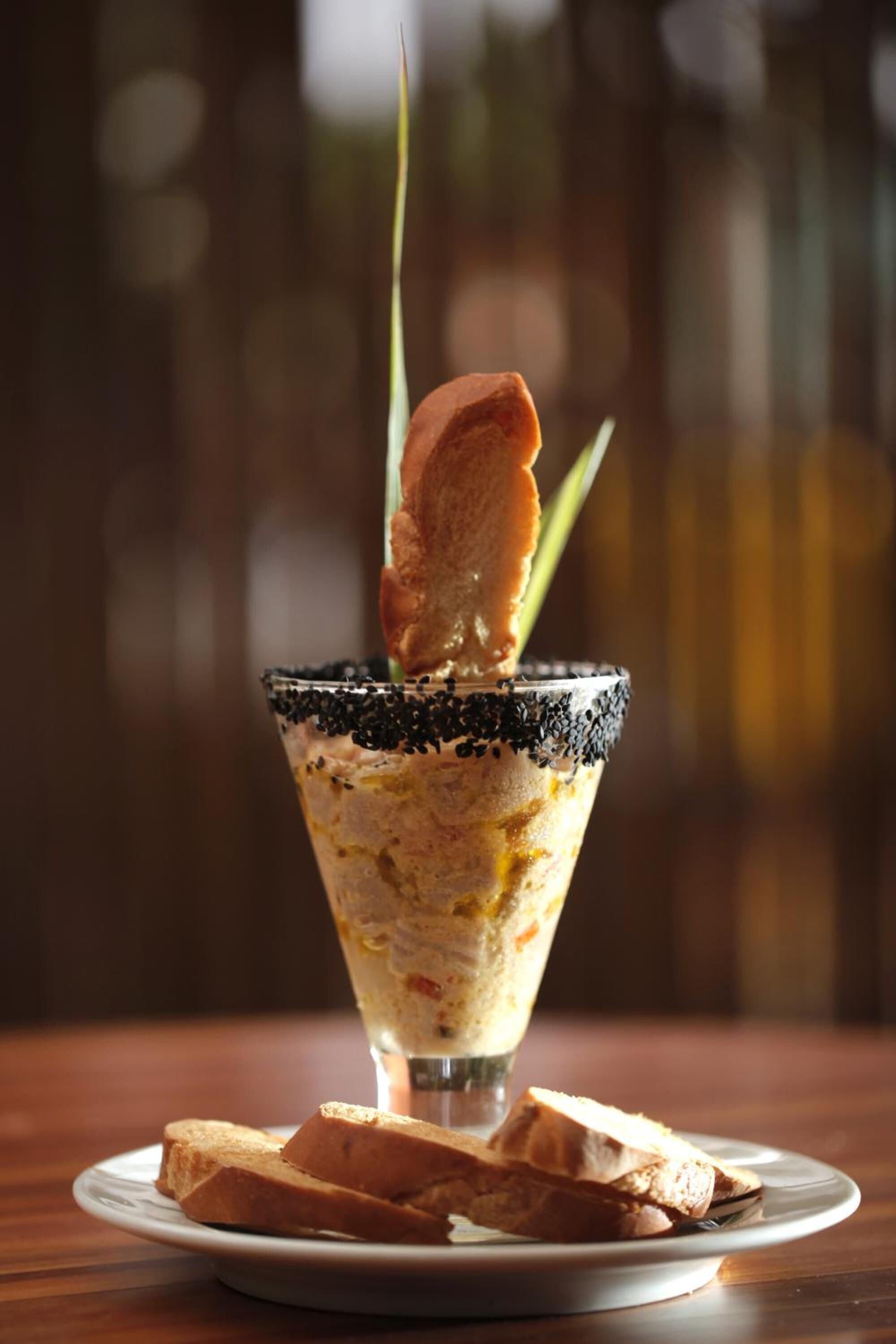 A cocktail in a martini glass with a black rim, garnished with a slice of orange and a green leaf, on a wooden table with a plate of bread.