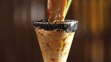A cocktail in a martini glass with a black rim, garnished with a slice of orange and a green leaf, on a wooden table with a plate of bread.