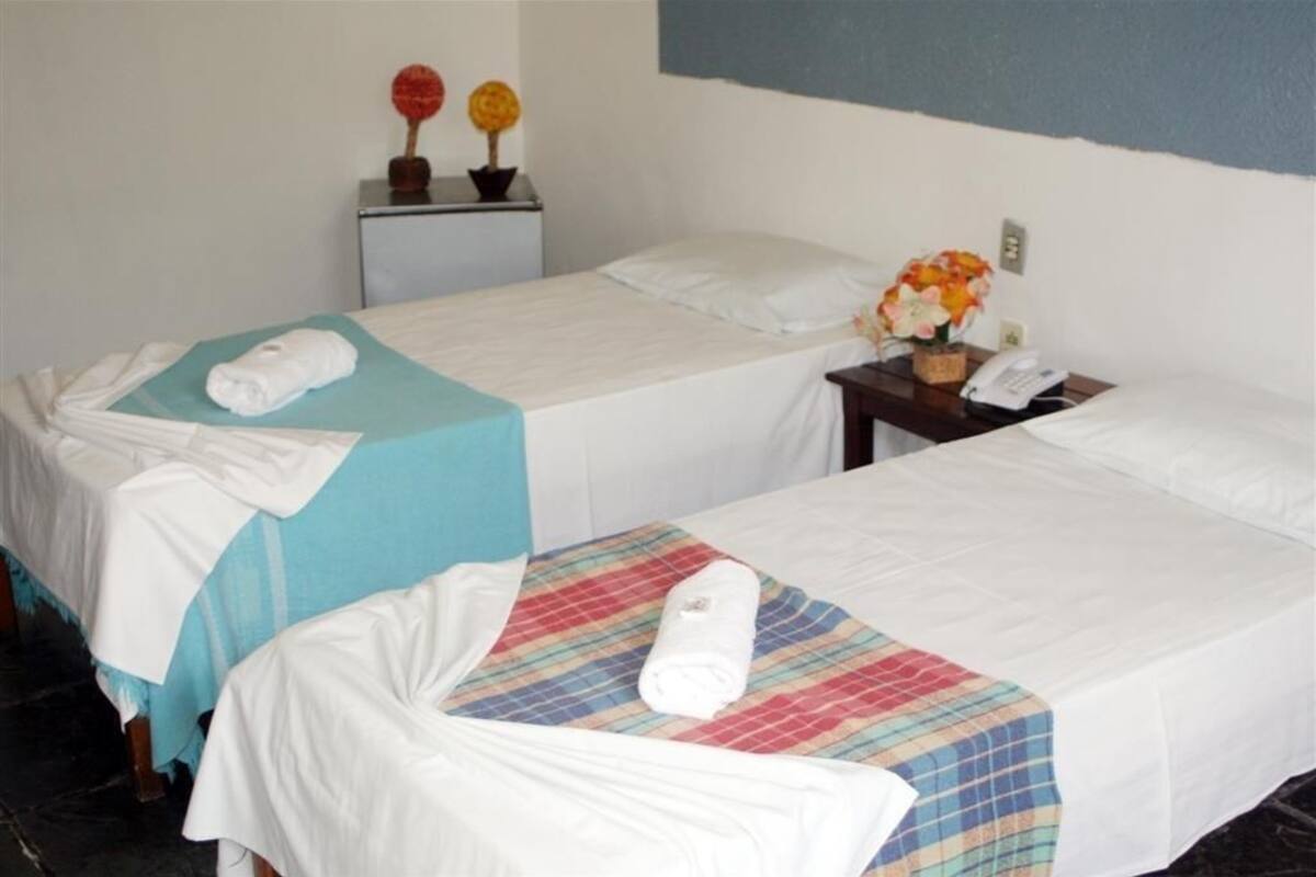 A hotel room with two single beds, each with a white bedspread and a rolled towel. There is a small table with a telephone and a vase of flowers.