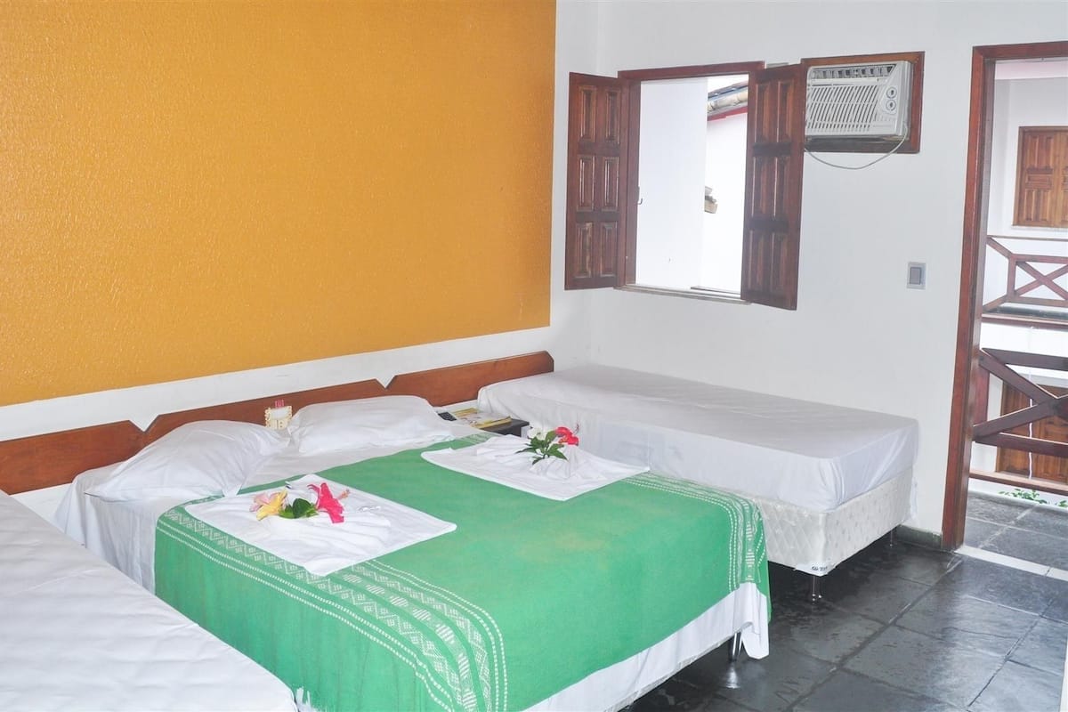 A hotel room with two beds, a green bedspread, white linens, a wooden headboard, a window with wooden shutters, and a small balcony with a wooden railing.