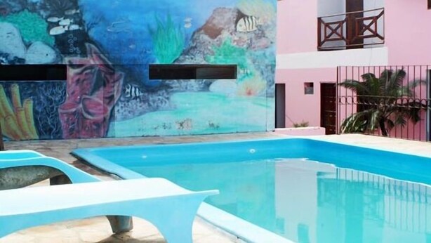 Une piscine ornée d’une fresque représentant un paysage sous-marin, un bâtiment rose doté de balcons et une clôture en métal.