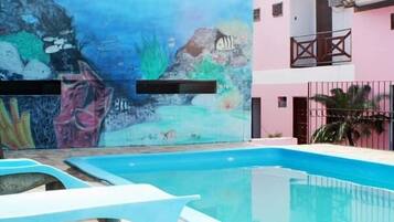 Una piscina con un mural de escena submarina, un edificio rosa con balcones y una cerca de metal.