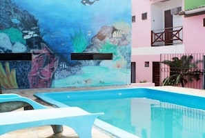 Una piscina con un mural de escena submarina, un edificio rosa con balcones y una cerca de metal.