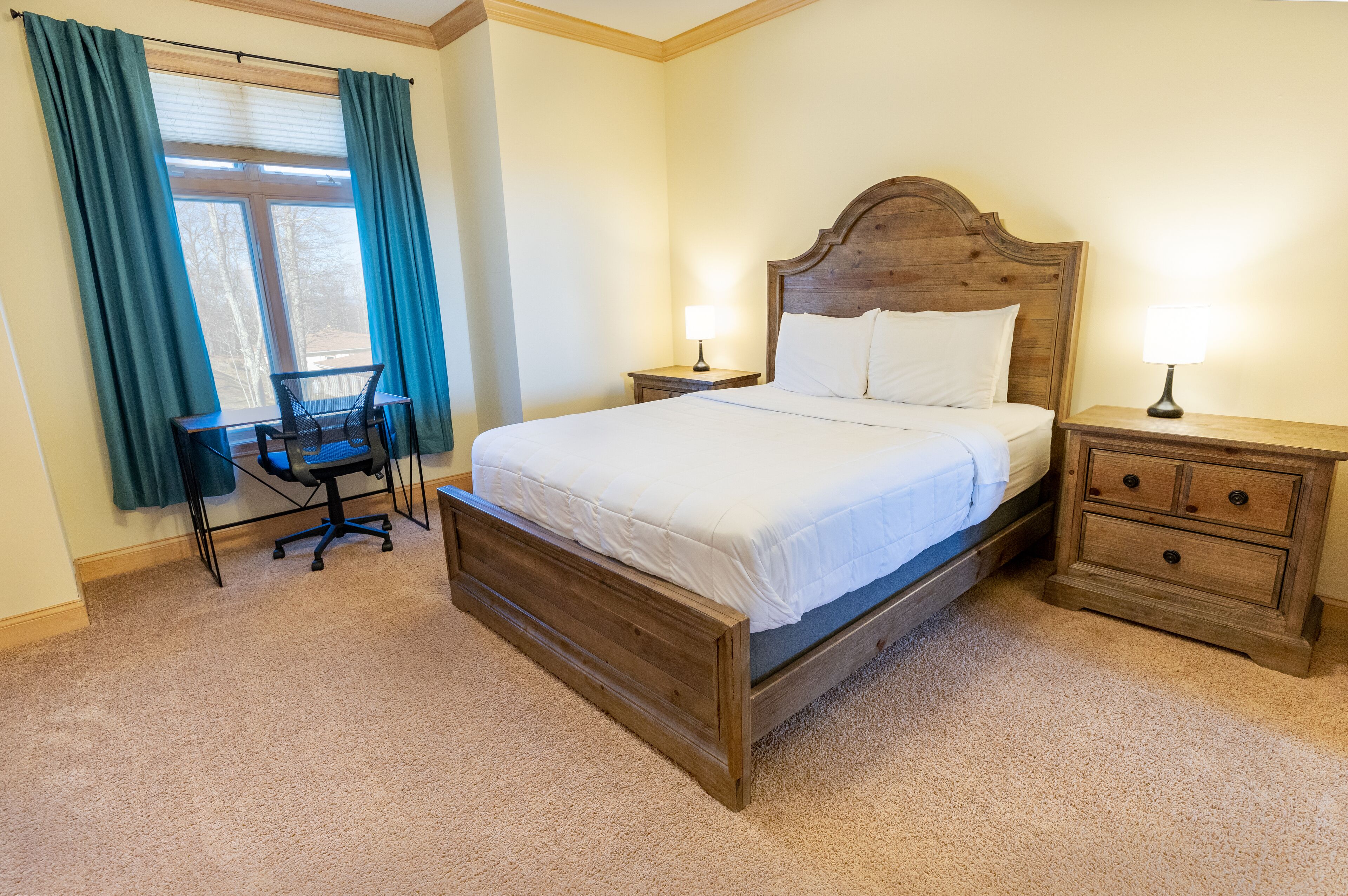 A bedroom with a wooden bed, two bedside tables with lamps, a desk with a chair, and a window with curtains.