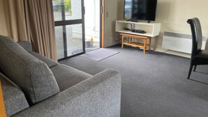 A living room with a grey sofa, a television on a white stand, and a black chair. - Allan Court Motel (Dunedin)