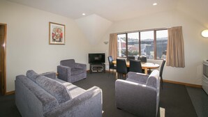 A modern living room with grey sofas, a flat-screen TV, a dining table with chairs, and a large window with curtains. - Allan Court Motel (Dunedin)