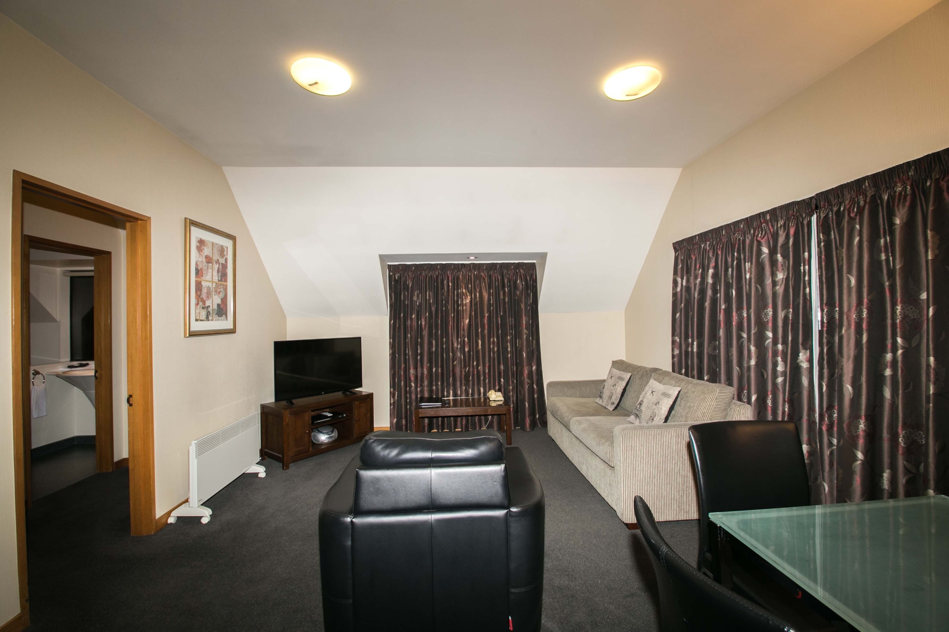 A living room with a sofa, a glass table, a black leather ottoman, a TV, and a radiator.