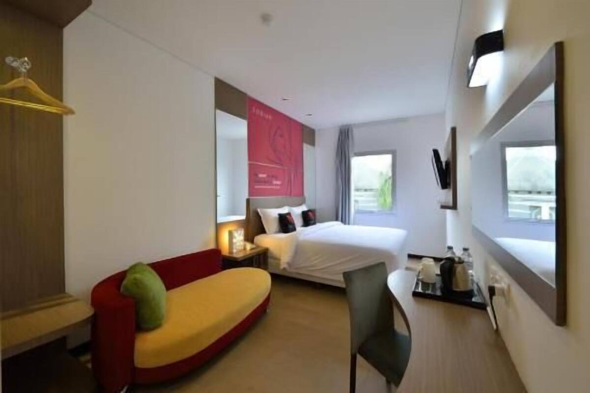 Une chambre d’hôtel avec un lit, un canapé rouge, un bureau avec une chaise, une télévision et une fenêtre donnant sur un paysage.