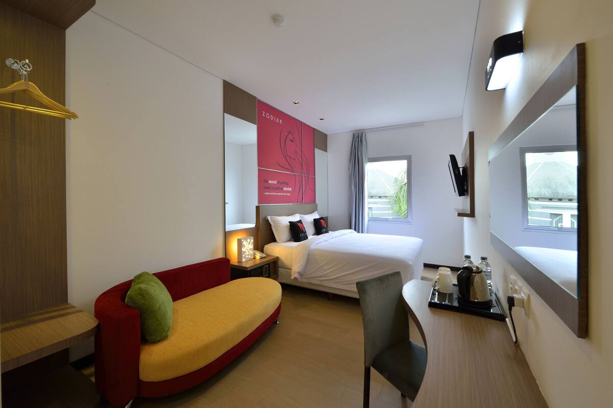 Habitación de hotel con cama, sofá rojo y amarillo, escritorio con silla, televisión y ventana con vistas.