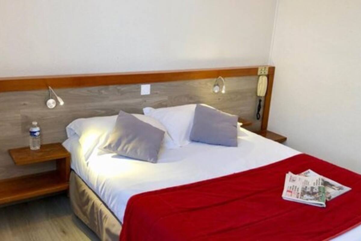 Une chambre d’hôtel avec une tête de lit en bois, un lit avec une literie blanche et deux oreillers, une table de chevet avec une bouteille d’eau, et un plaid rouge sur le lit.