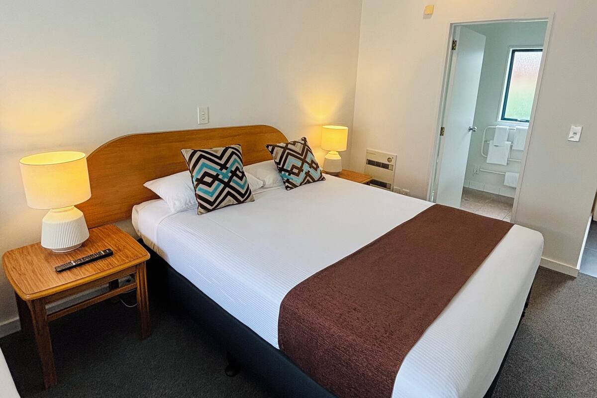 A neatly made bed with a brown blanket, two bedside lamps, and a remote control on a wooden nightstand.