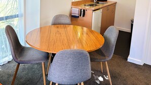 A compact living space with a round wooden dining table surrounded by four grey chairs, a kitchenette with a microwave and a kettle, and a window with a view of greenery.