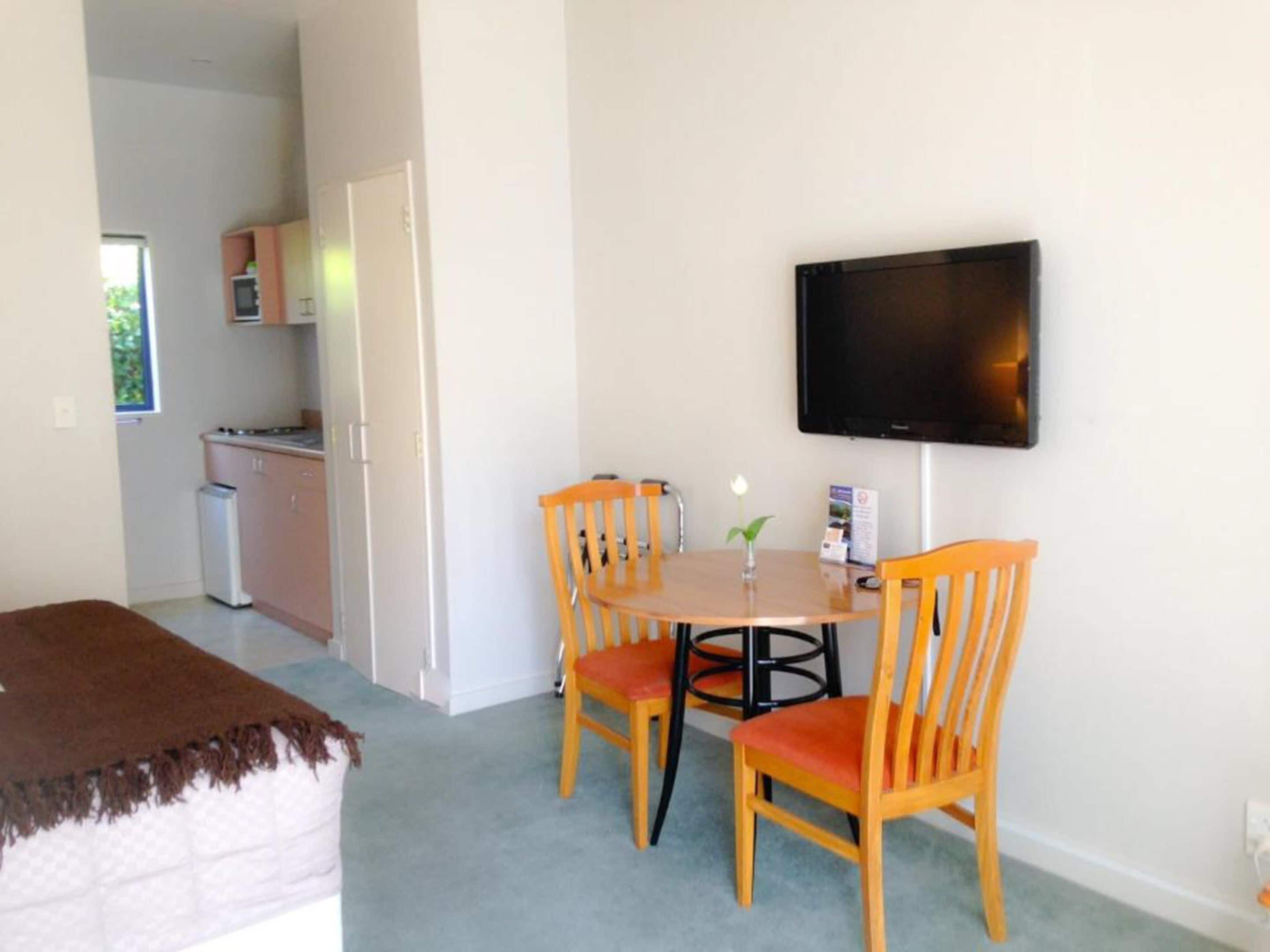 Una habitación pequeña y acogedora con una cama, una mesa de comedor con cuatro sillas, un televisor en la pared y una kitchenette al fondo.