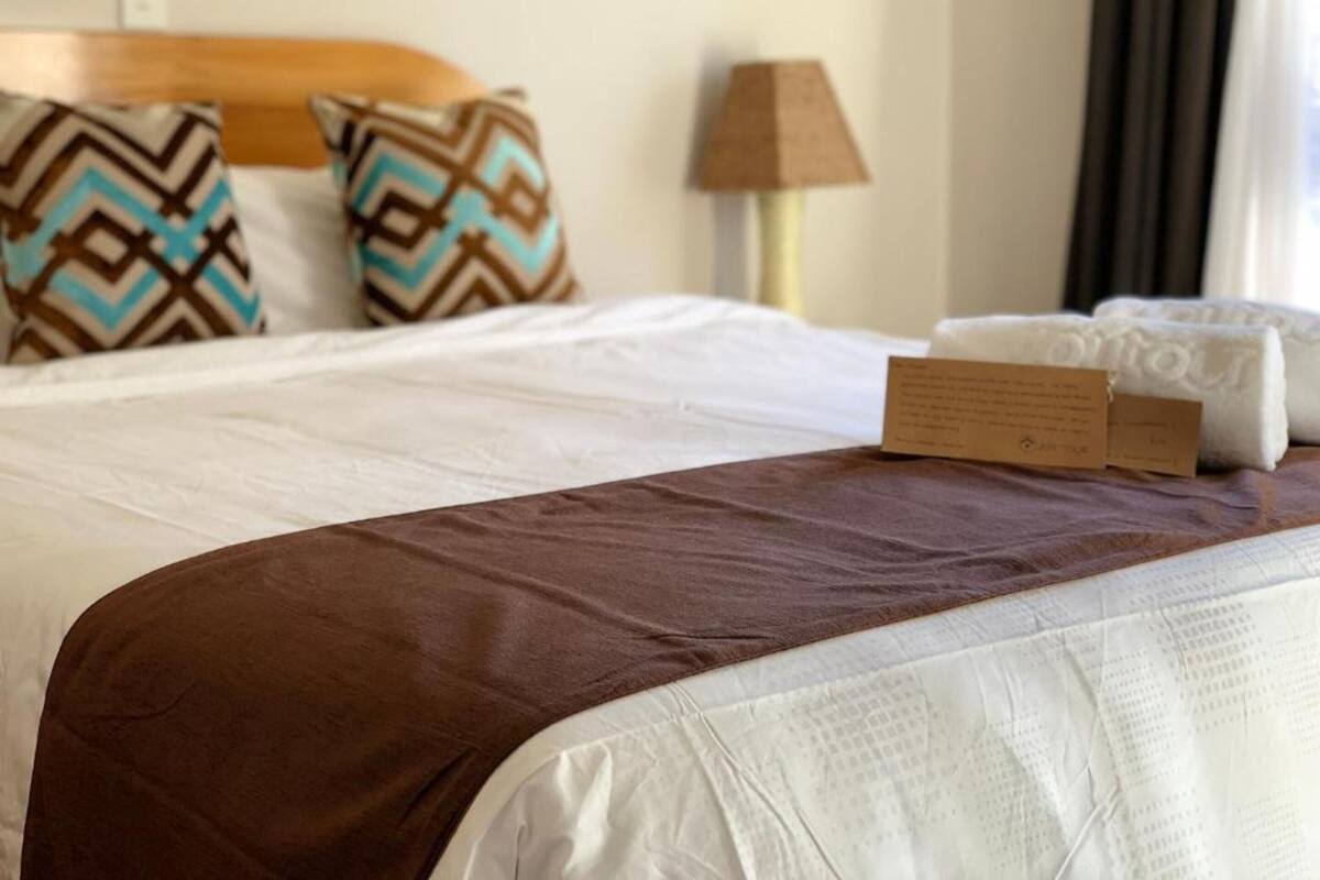 A neatly made bed with a brown bedspread, white linens, and patterned pillows. There is a lamp and a sign on the side of the bed.