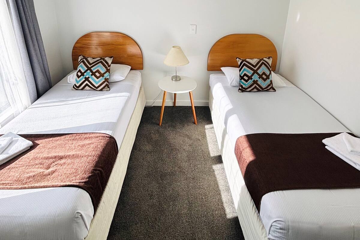 A hotel room with two single beds, a small bedside table with a lamp, and a carpeted floor.
