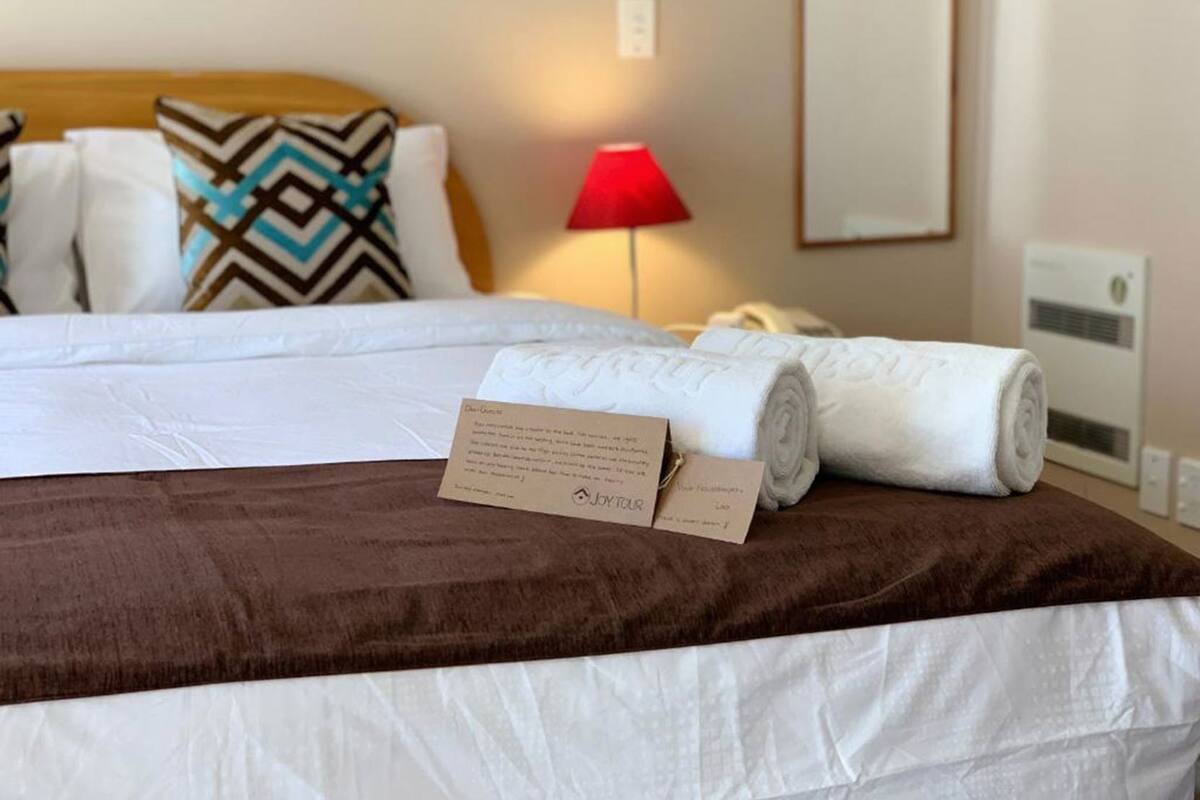 A neatly made bed with a brown bedspread, white pillows, and a rolled white towel. There is a red lamp on the nightstand, a card on the bed, and a wall-mounted air conditioner.