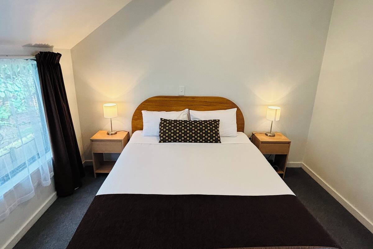 A neatly made bed with white and brown bedding, two bedside lamps, and a window with curtains.