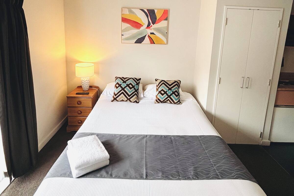 A neatly made bed with a grey and white duvet, two patterned pillows, a white towel, and a bedside lamp. There is a wardrobe with a mirror and a colorful abstract painting on the wall above the bed.