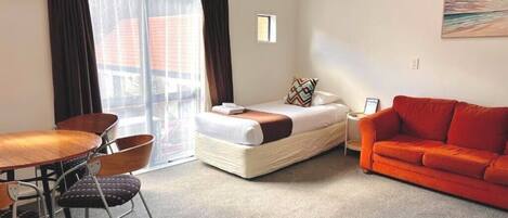 Una habitación de hotel con cama, sofá rojo, mesa de comedor con sillas y una ventana pequeña.