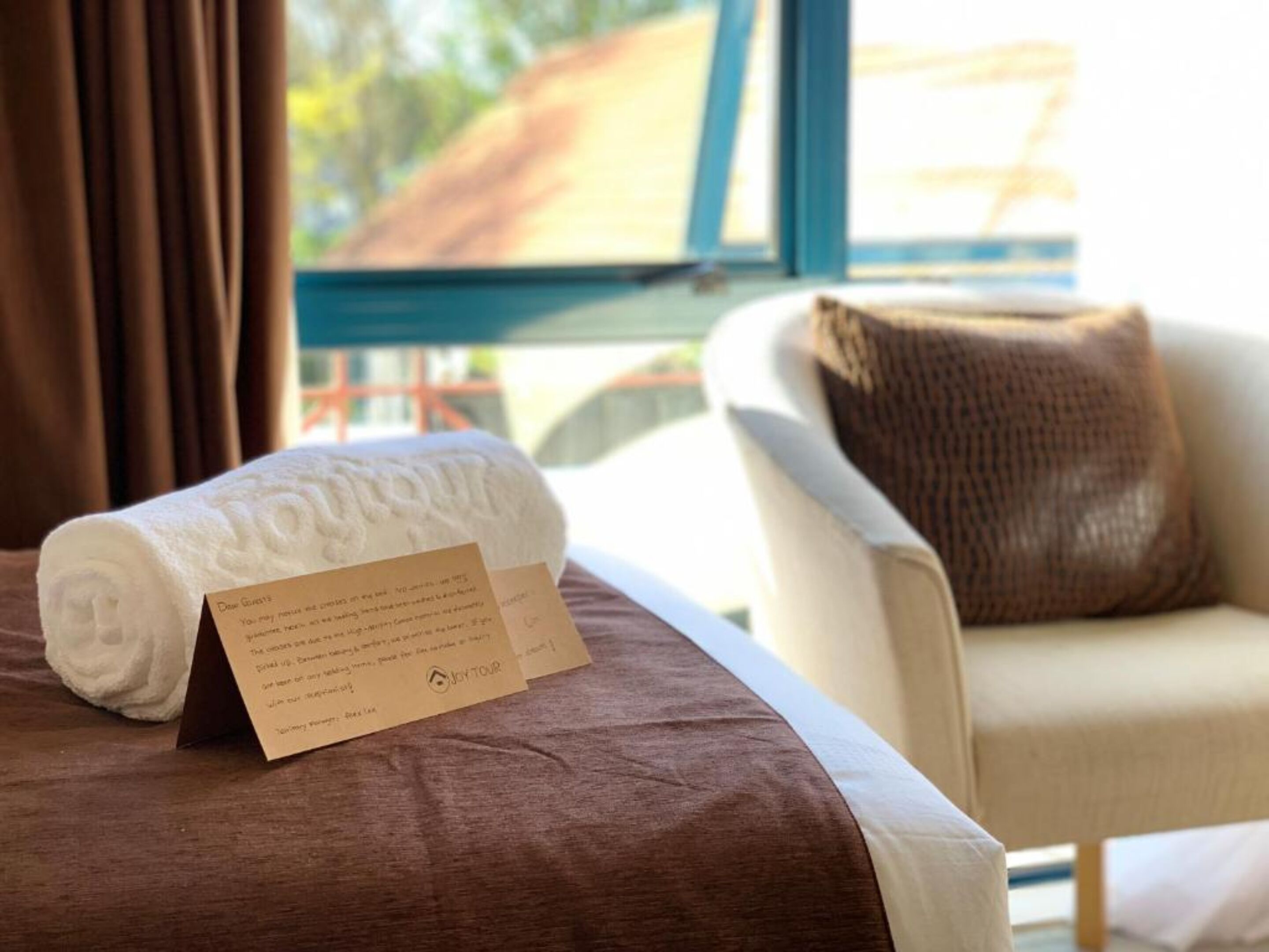 A neatly made bed with a folded towel and a welcome card on a wooden floor.