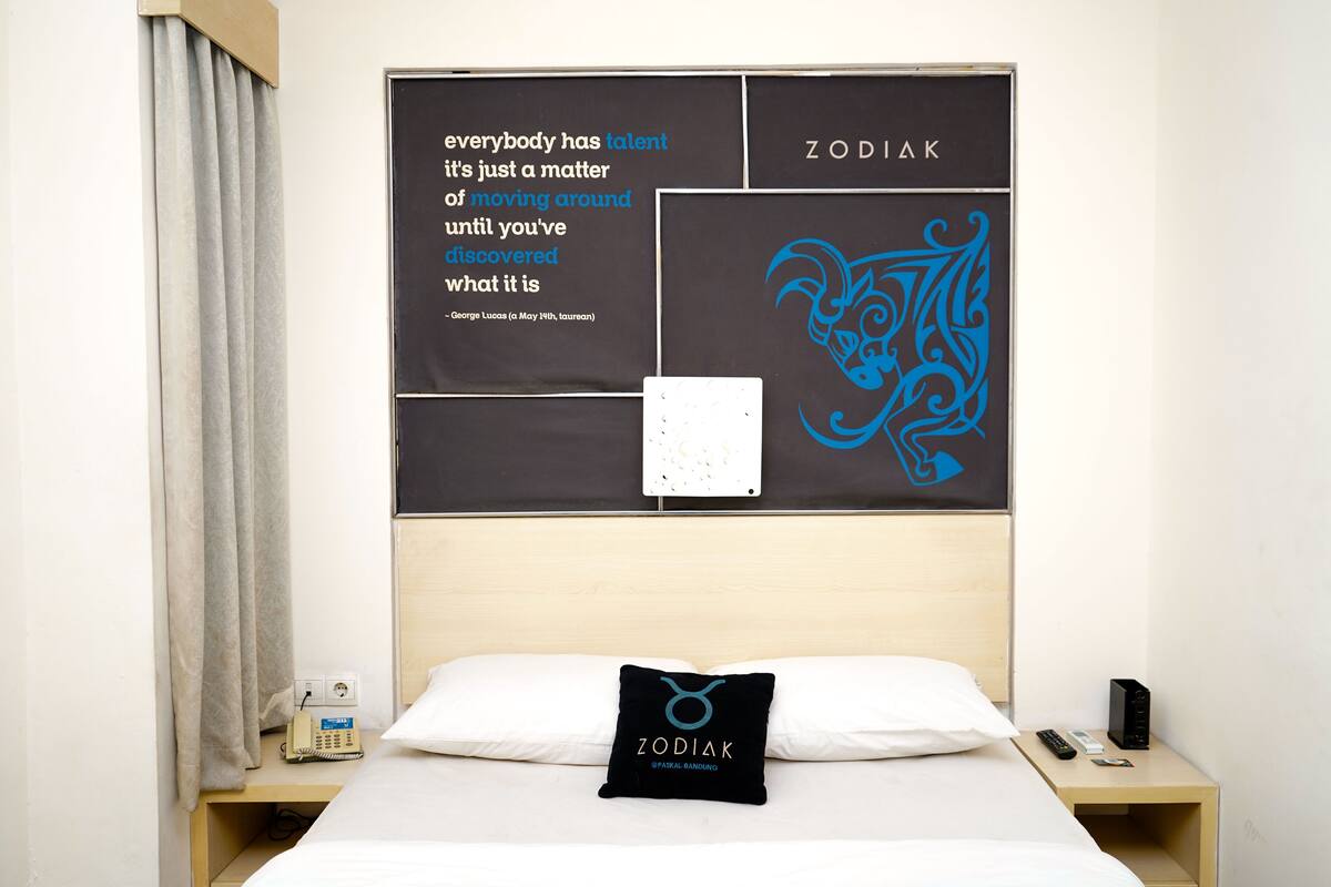 Ett hotellrum med en säng, en kudde med en symbol för ett stjärntecken, en vägg med ett citat och ett stjärnteckensymbol, och ett nattduksbord med en telefon och en fjärrkontroll.