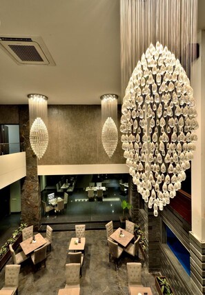 A restaurant with a high ceiling, two chandeliers, and a large, decorative wall hanging. Tables and chairs are arranged for dining. - Serela Merdeka Bandung (Bandung)