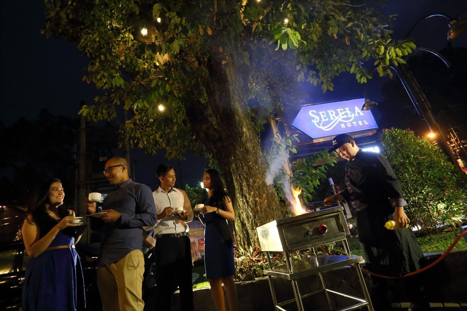 Un grupo de personas cenando al aire libre por la noche, con un letrero del 'Serena Hotel' al fondo.