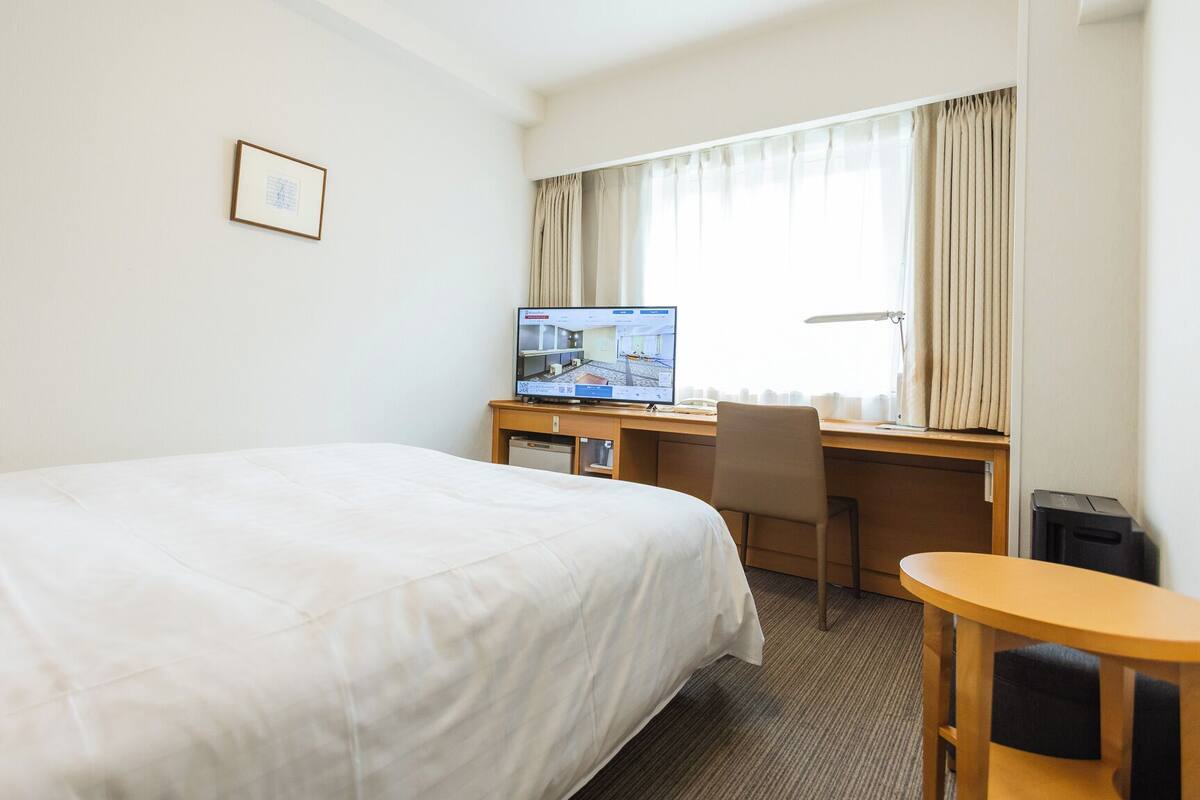 A hotel room with a bed, desk, chair, computer, and window with curtains.