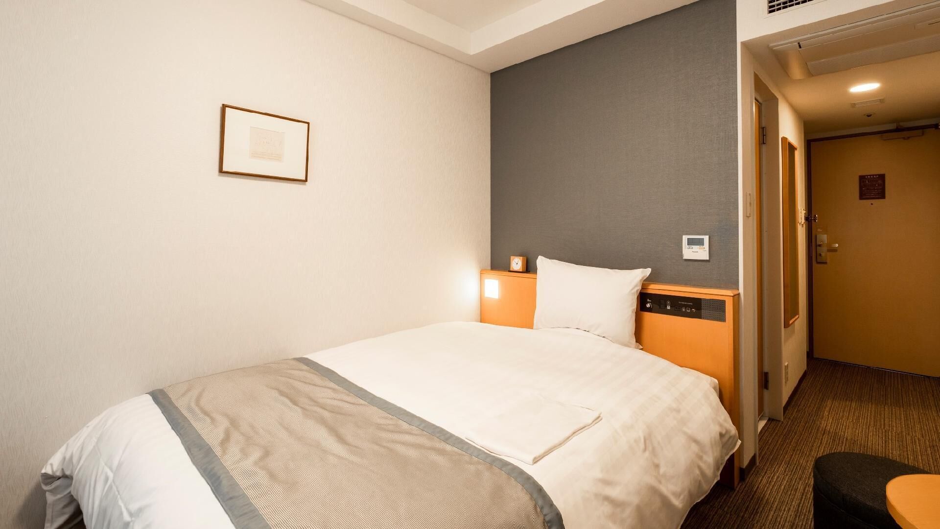A hotel room with a bed, a nightstand, a wall clock, a thermostat, and a door.
