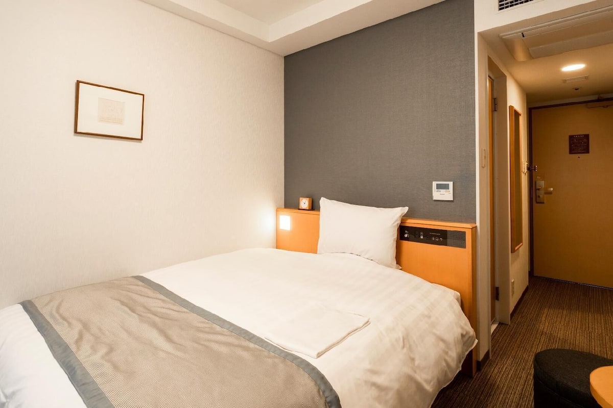A hotel room with a bed, a nightstand, a wall clock, a thermostat, and a door.
